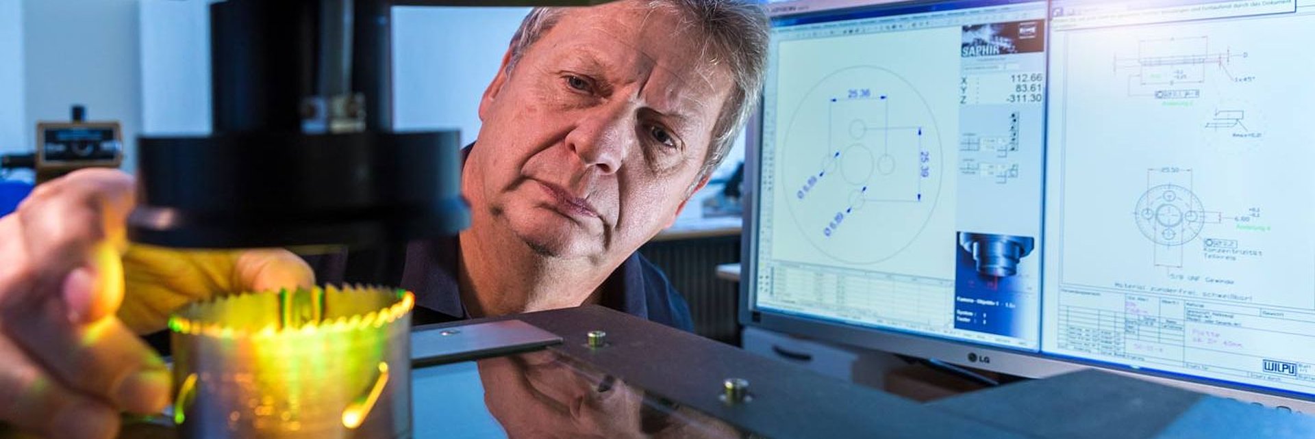 Mann fokussiert auf Lochsäge neben Computerbildschirmen mit technischen Zeichnungen.